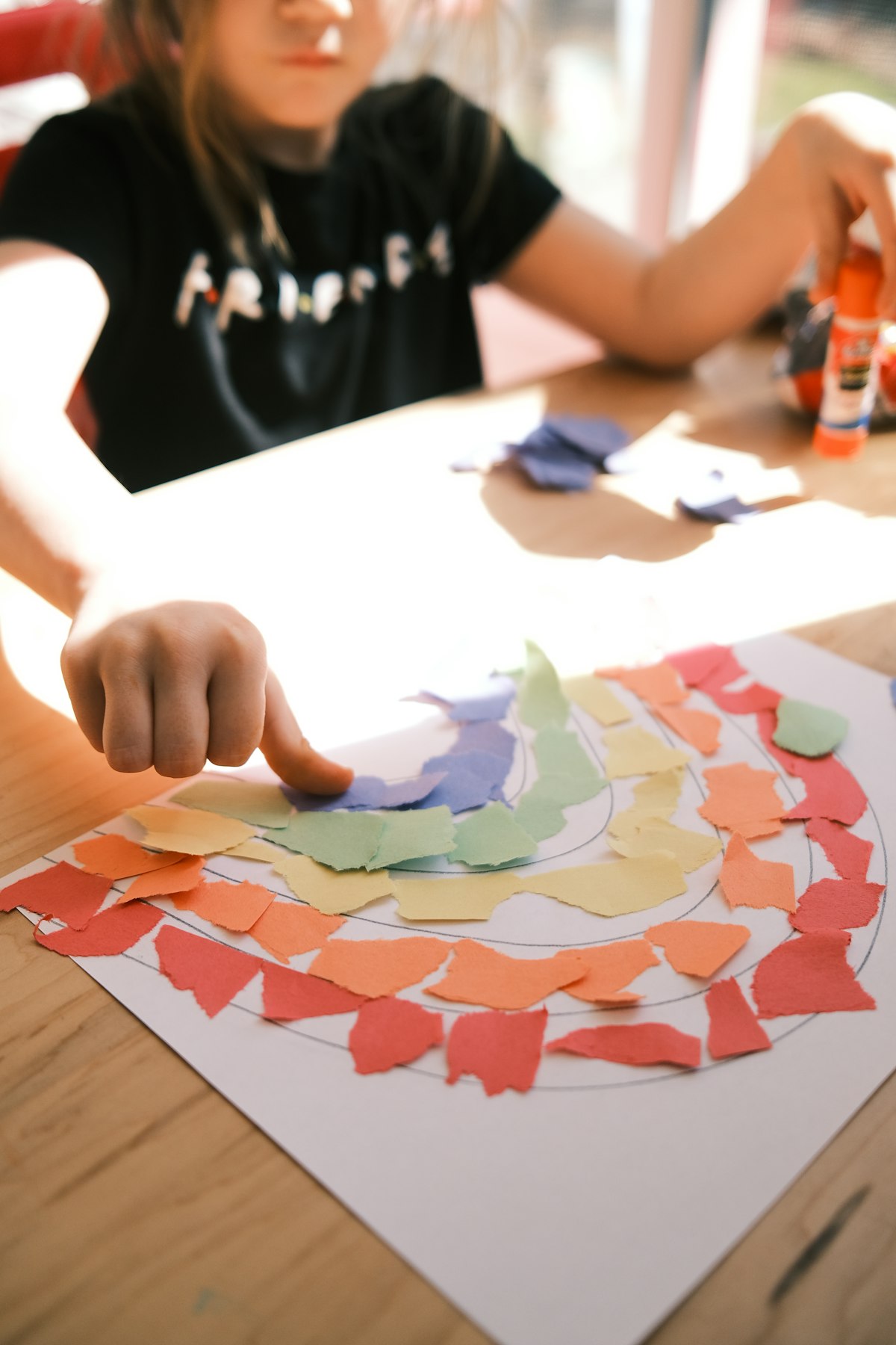 A child gluing colourful paper scraps onto a rainbow drawing